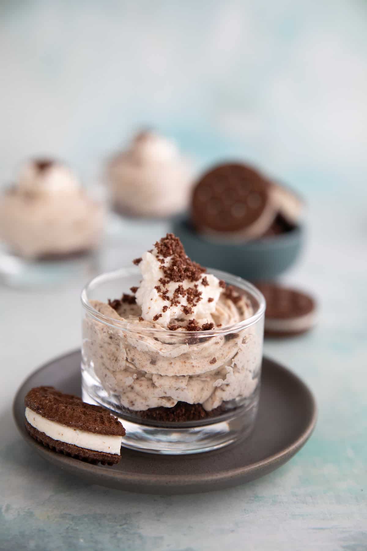A cup of Keto Cookies and Cream Mousse sits on a plate with broken keto cookies around it.