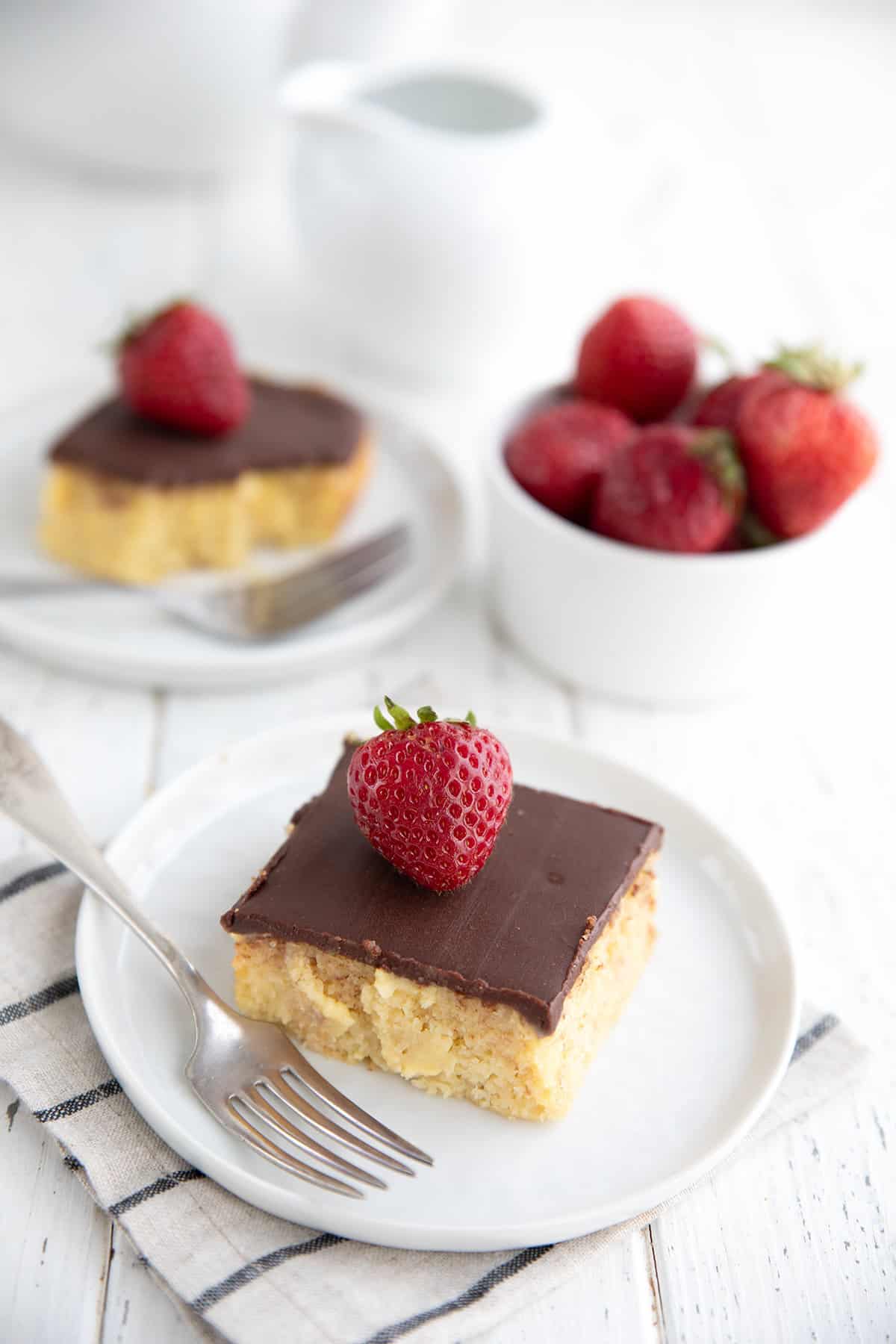 Two slices of Keto Boston Cream Pie Poke Cake sit on a white wooden table with a bowl of strawberries.