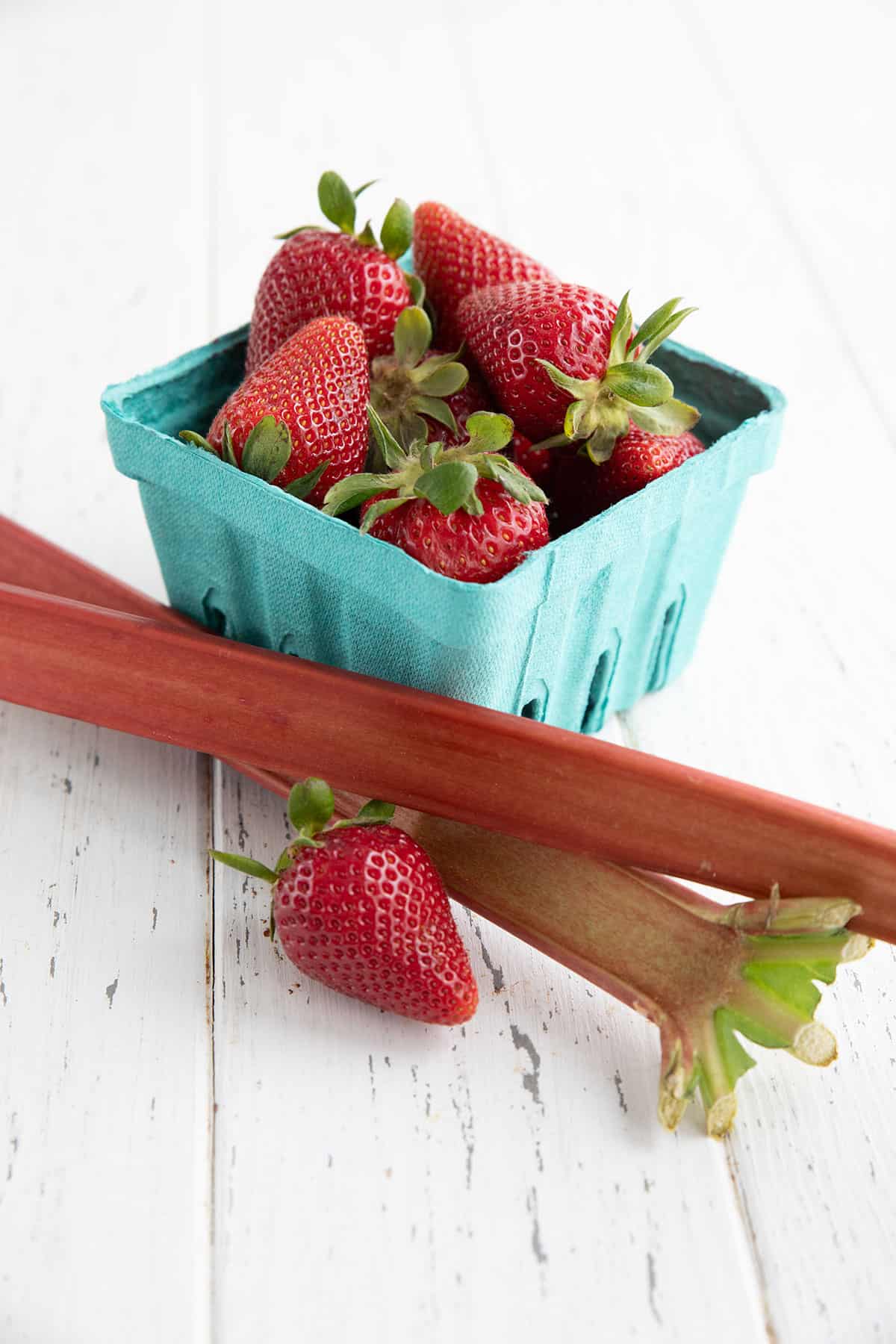 A container of fresh strawberries and some rhubarb stalks on a white wooden table.