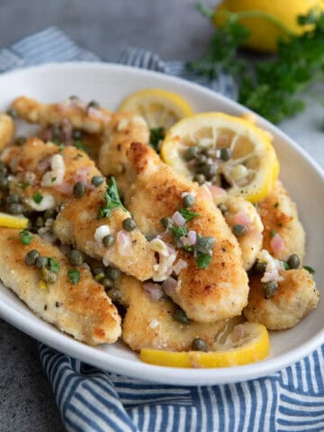 Pieces of chicken piccata with sauce in a white oval serving dish over a blue patterned napkin.