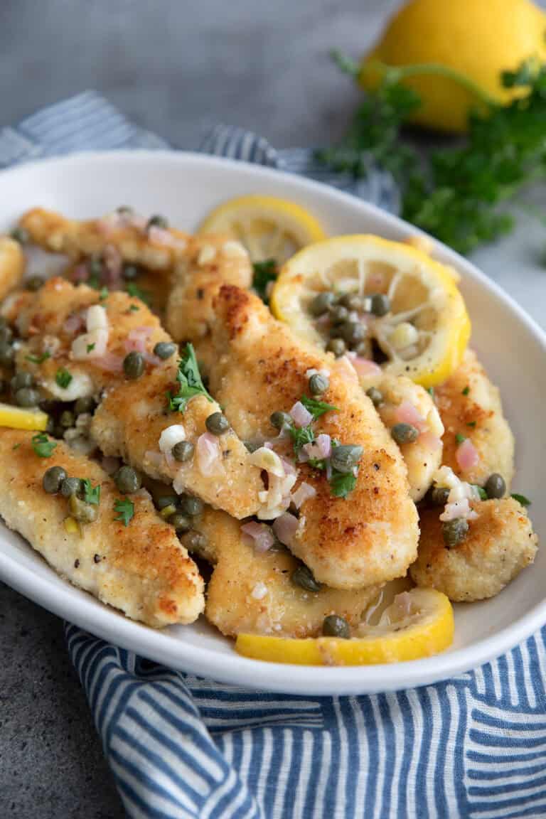 Pieces of chicken piccata with sauce in a white oval serving dish over a blue patterned napkin.