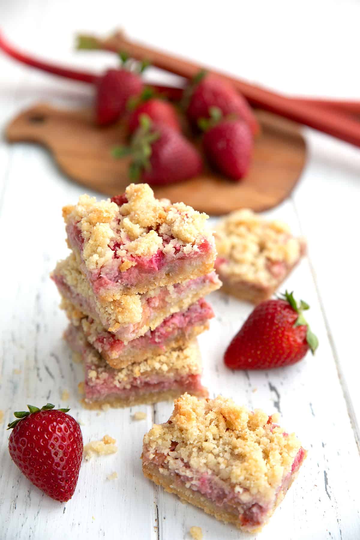 A stack of strawberry rhubarb crumb bars on a white table, with berries and rhubarb on a cutting board in the background.