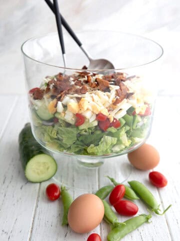 7 Layer Salad arranged in a trifle dish with cucumber, peas, tomatoes, and eggs on the table.