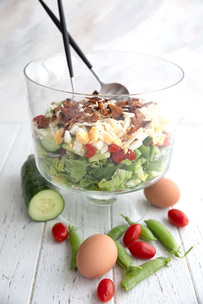 7 Layer Salad arranged in a trifle dish with cucumber, peas, tomatoes, and eggs on the table.