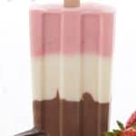 Titled Pinterest image of a keto neapolitan popsicle on a white table with strawberries and chocolate around it.