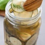 Titled image of a jar of spicy refrigerator pickles, with wooden tongs lifting one out of the jar.