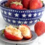 Two Cheesecake Stuffed Strawberries with graham cracker crumbs on a small pewter plate in front of a bowl of strawberries.