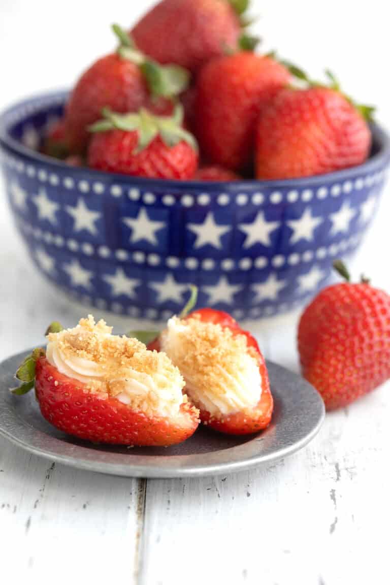 Two Cheesecake Stuffed Strawberries with graham cracker crumbs on a small pewter plate in front of a bowl of strawberries.