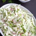 Titled image of Dill Pickle Coleslaw in an oval dish with pickles in the background.