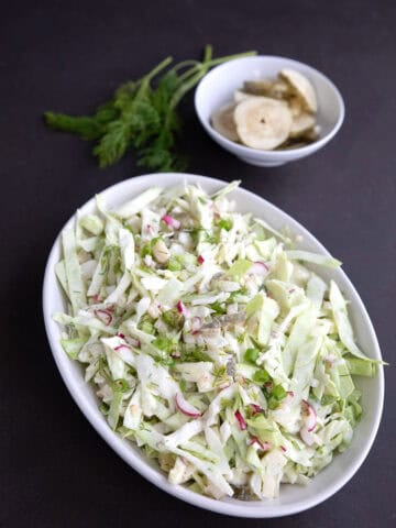 Dill Pickle Keto Coleslaw in a white oval dish with a bowl of pickles and some fresh dill in the background.