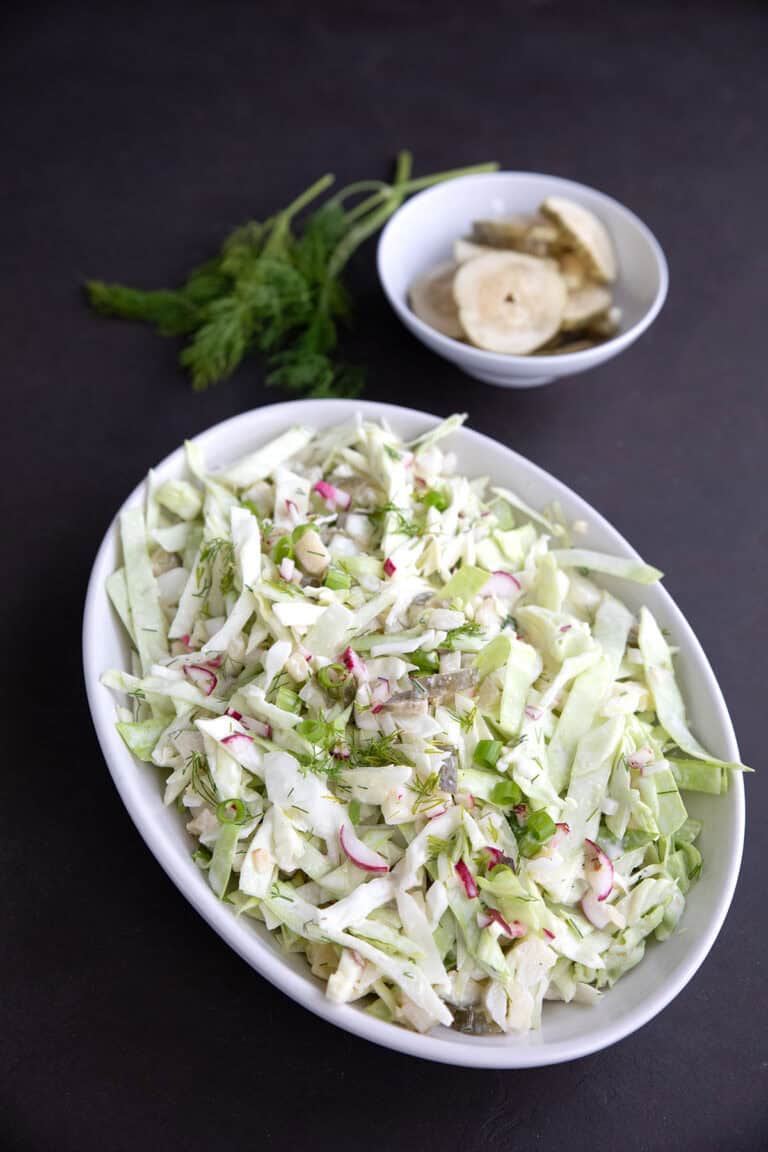 Dill Pickle Keto Coleslaw in a white oval dish with a bowl of pickles and some fresh dill in the background.