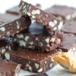 Close up shot of keto chocolate crunch bars cut up on a white table with chunks of sugar-free chocolate and a spoonful of peanut butter.