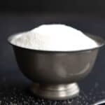 Titled image of a bowl of erythritol sweetener on a black background with a spoon of sweetener in front.