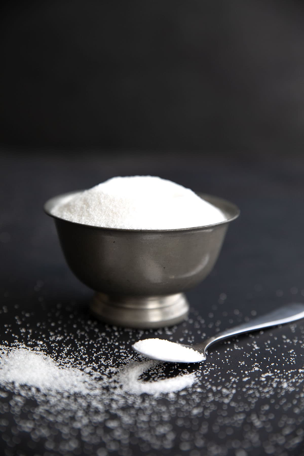 A metal bowl of erythritol sweetener on a black background with a spoon of sweetener in front.