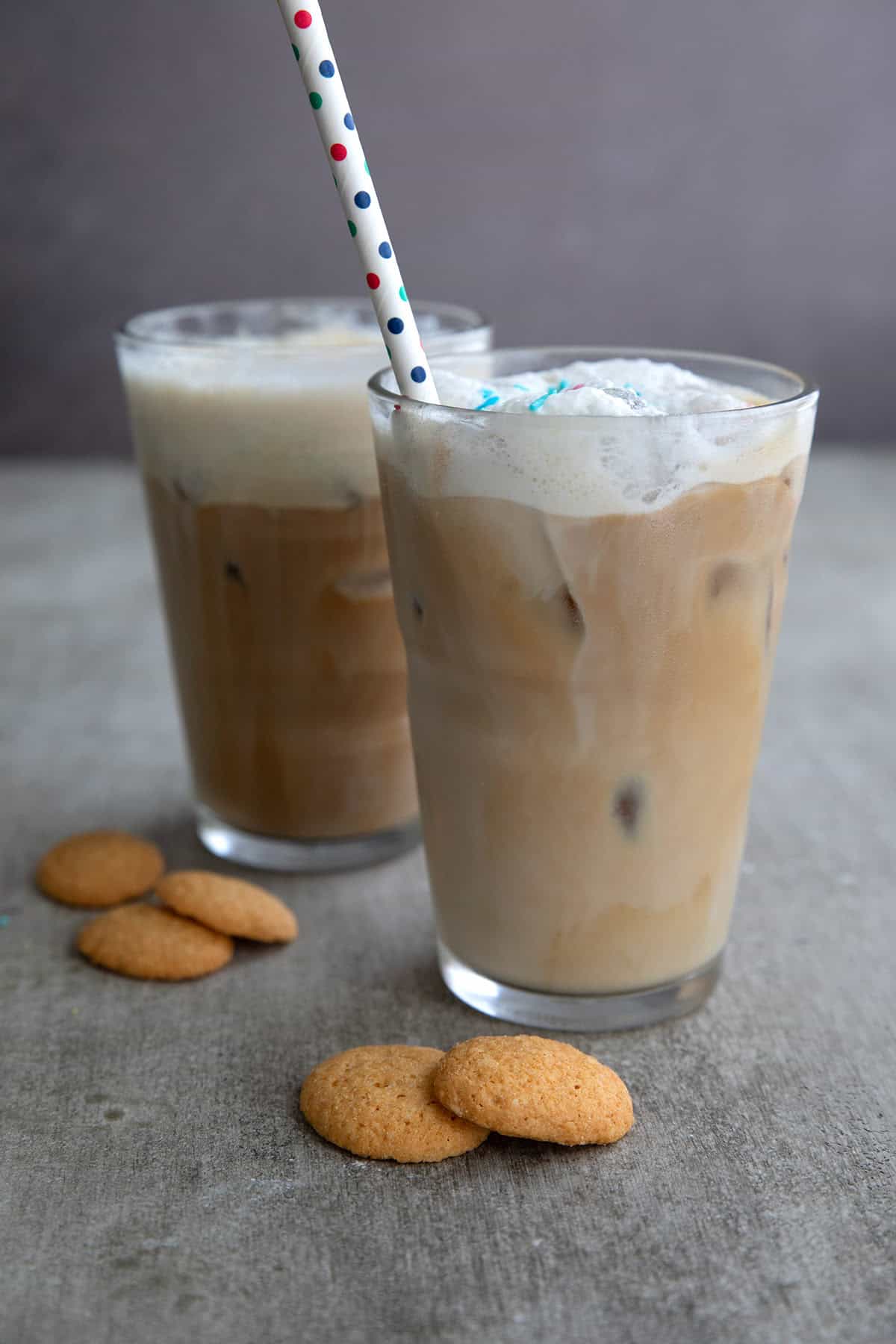 Two Keto Sugar Cookie Iced Lattes on a gray table with a polka dot straw in the front glass. Some small keto cookies sit near the glasses.