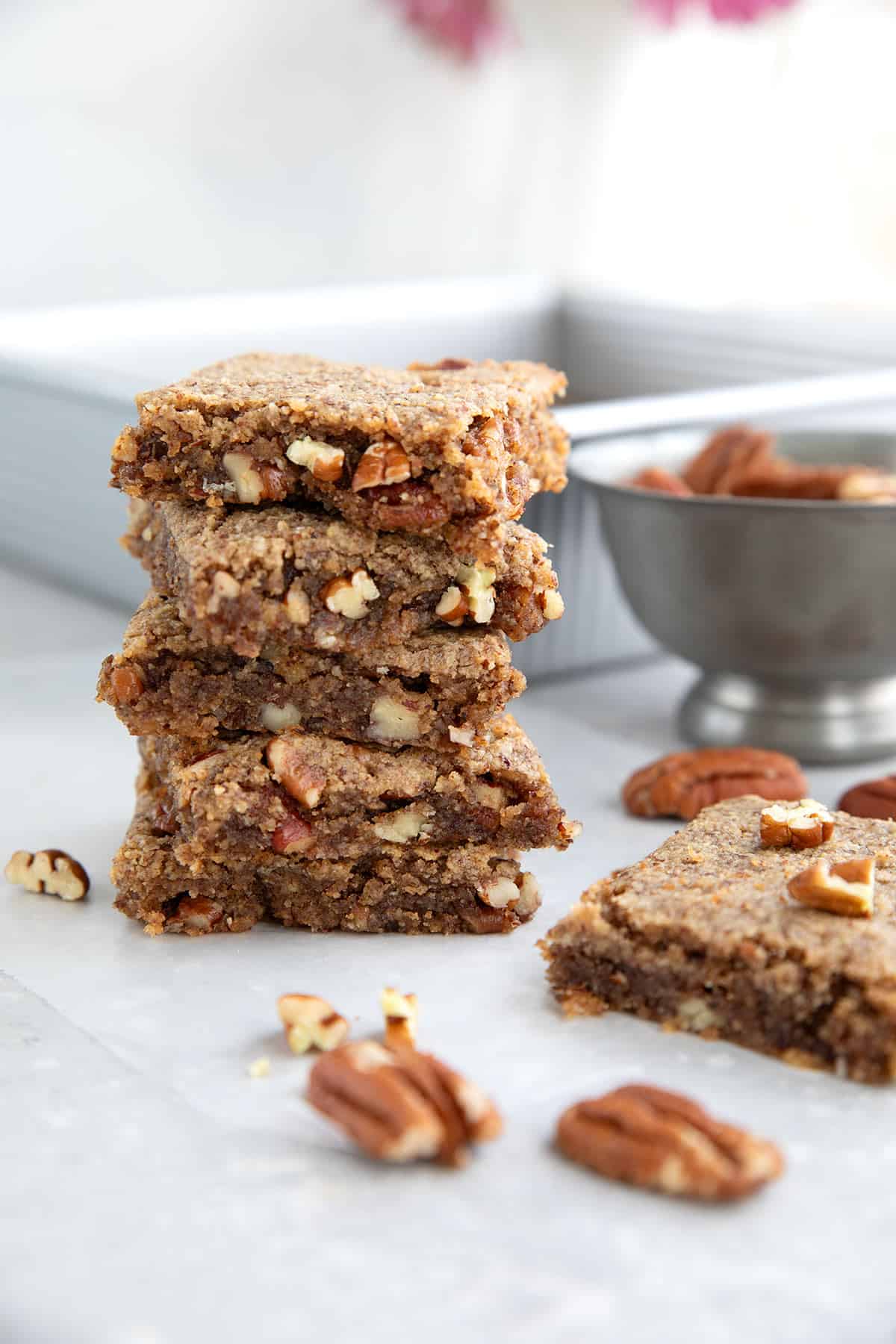 A stack of Keto Pecan Blondies on a gray table in front of the baking pan with more blondies.