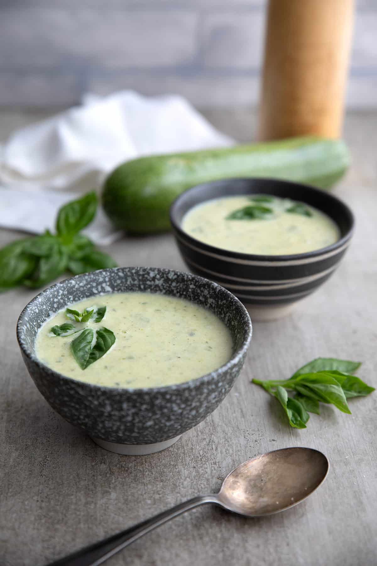 Two gray bowls of zucchini soup with zucchini and a pepper grinder in the background.