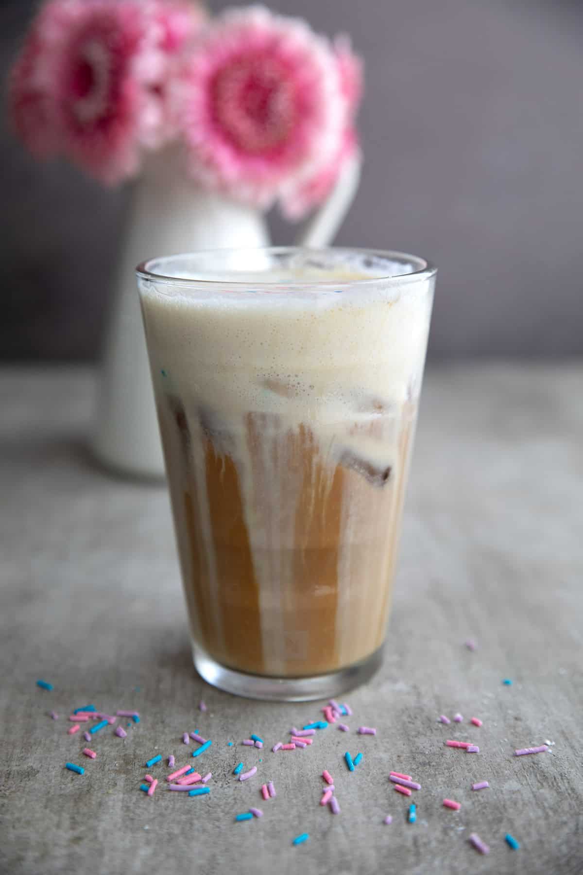 A glass of protein coffee on a table with a vase of pink flowers in the background.