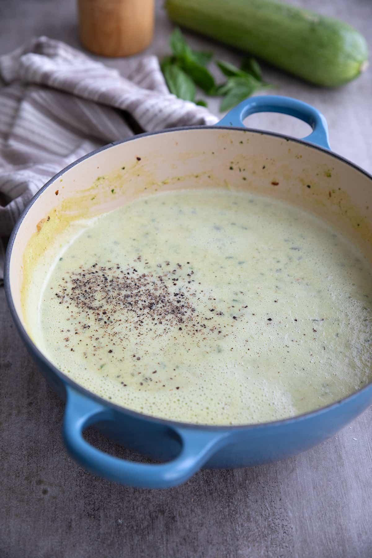 A blue dutch oven filled with creamy zucchini soup sprinkled with pepper.