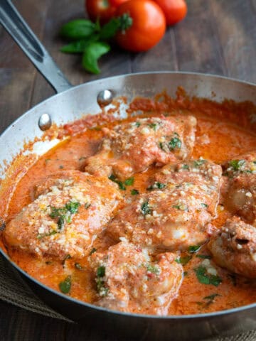 Tomato basil chicken in a skillet on a wooden table, with tomatoes and basil in the background.