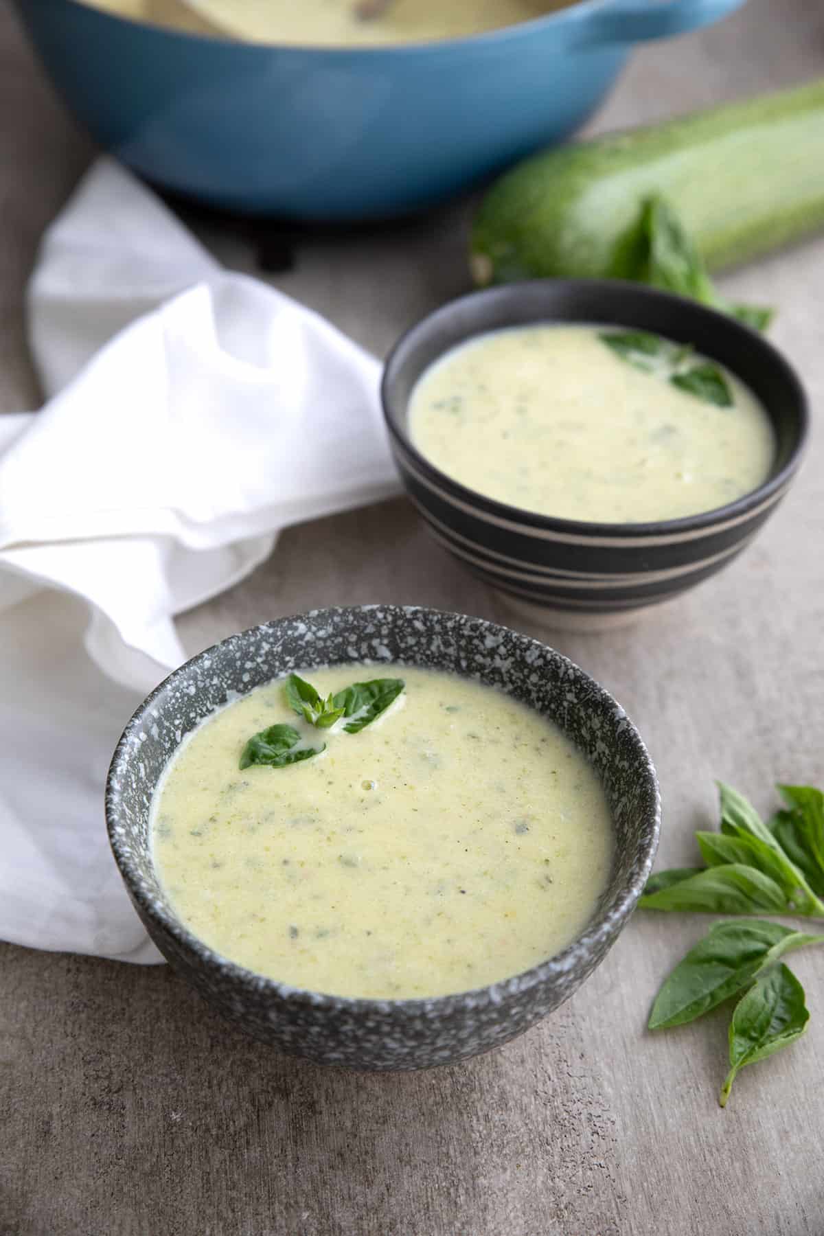 Two dark colored bowls of zucchini soup with a white napkin on a gray table.