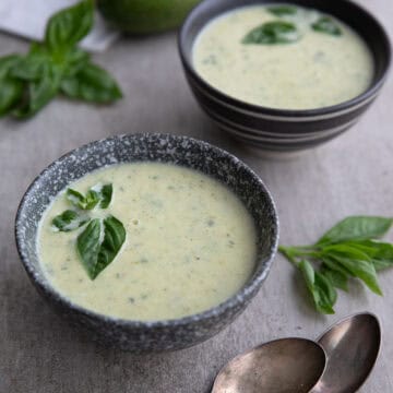 Two gray bowls of creamy zucchini soup on a concrete table with tarnished silver spoons.