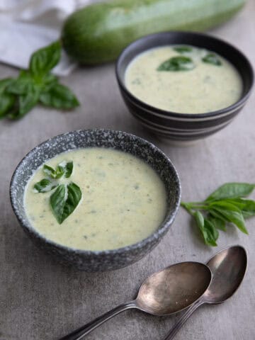 Two gray bowls of creamy zucchini soup on a concrete table with tarnished silver spoons.