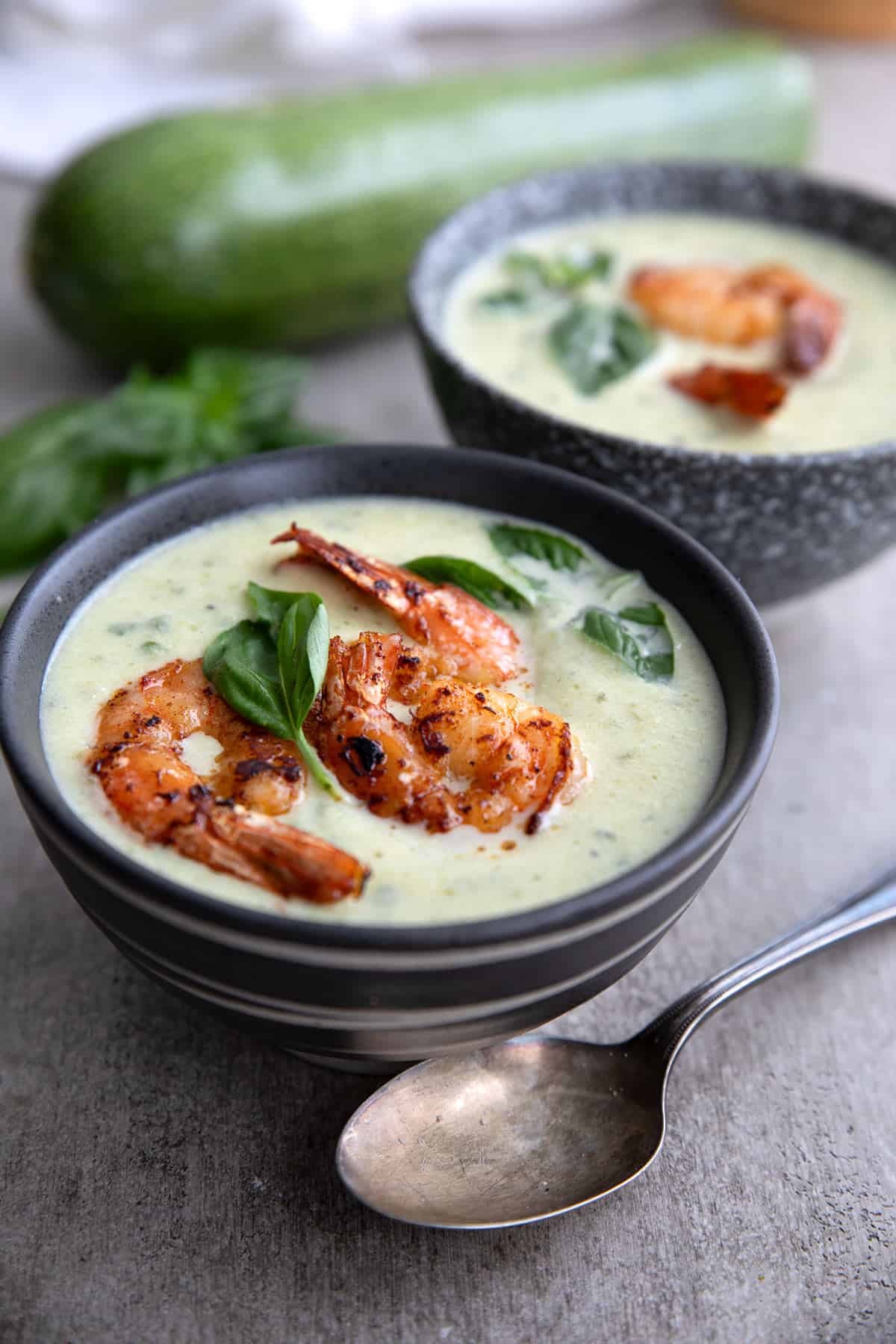 Two bowls of zucchini soup with spicy shrimp on a gray concrete table with zucchini in the background.