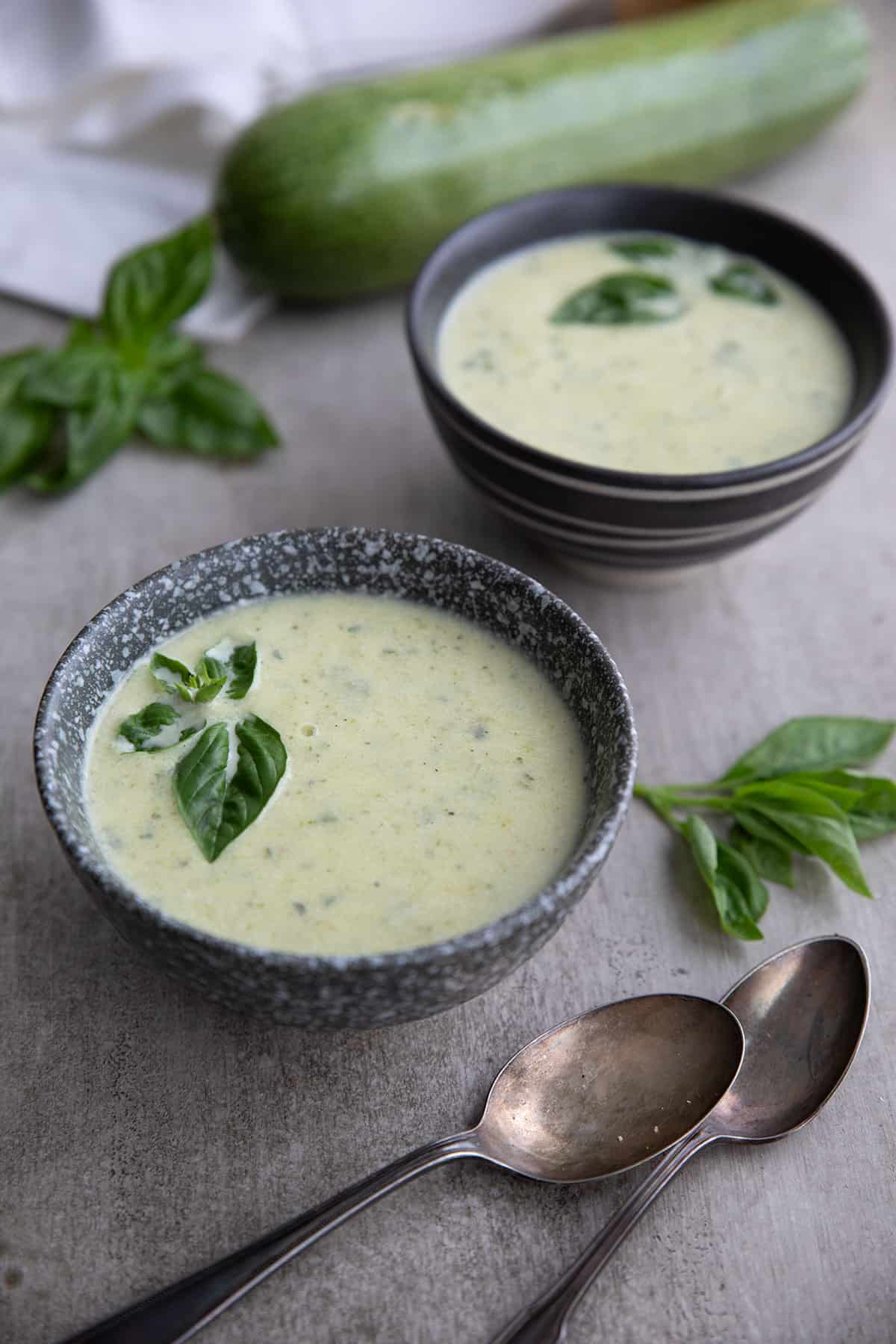 Two gray bowls of creamy zucchini soup on a concrete table with tarnished silver spoons.