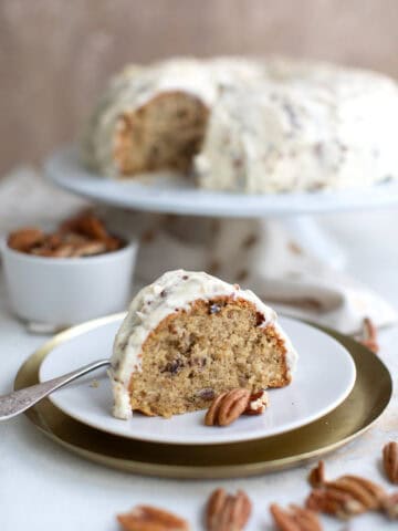 A slice of Keto Butter Pecan Pound Cake on a white plate, in front of the rest of the cake on a white cake stand.