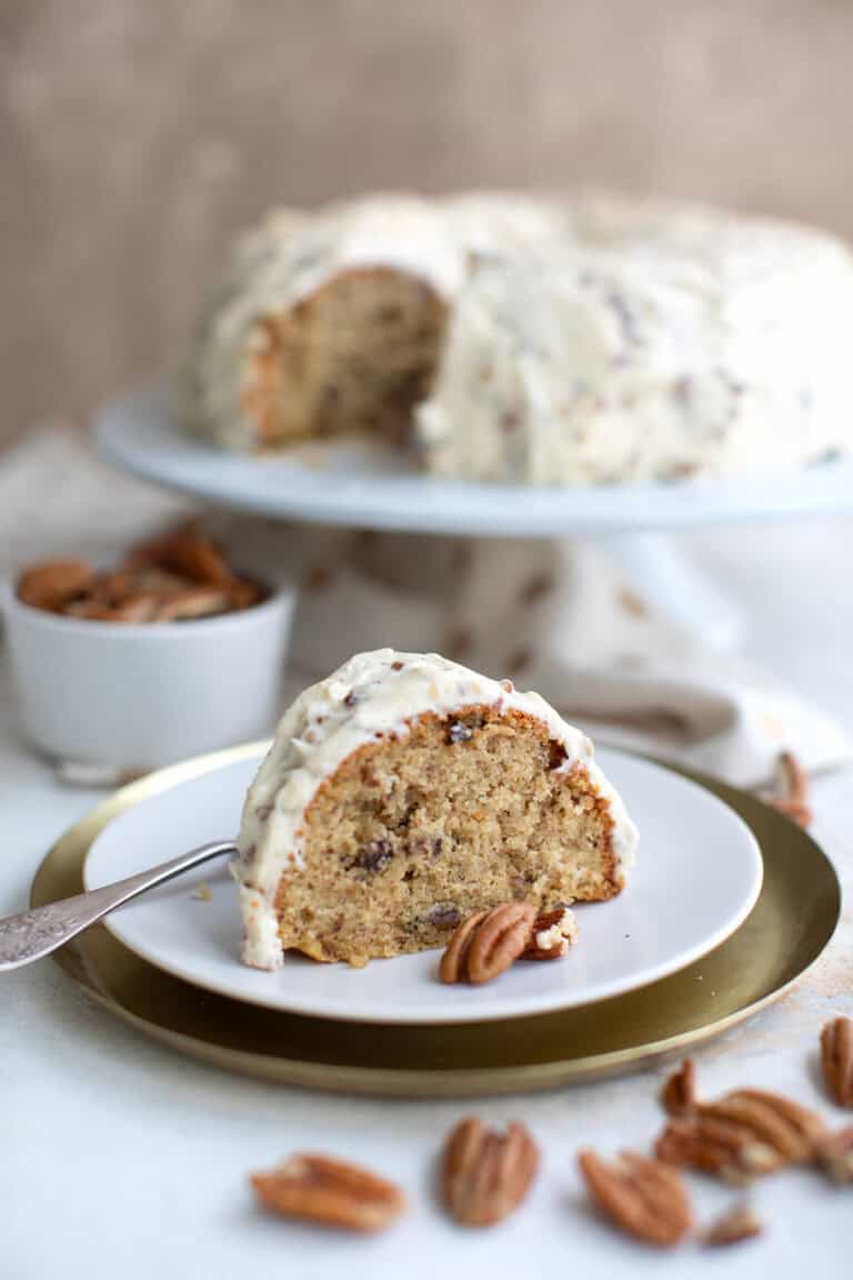 A slice of Keto Butter Pecan Pound Cake on a white plate, in front of the rest of the cake on a white cake stand.