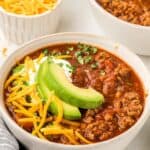 Titled image of a white bowl filled with Keto Chili, with slices of avocado and shredded cheese on top.