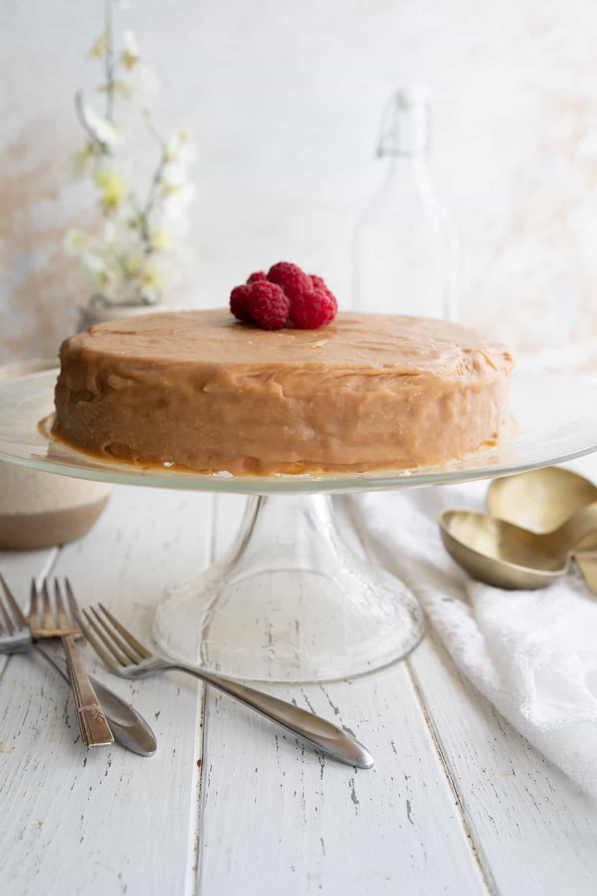 Keto Caramel Cake with raspberries on top, on a white table. Forks and measuring cups scattered around.