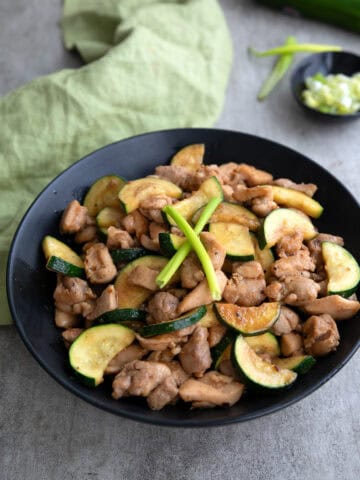 A black bowl filled with Chicken Zucchini Stir Fry on a gray concrete table.