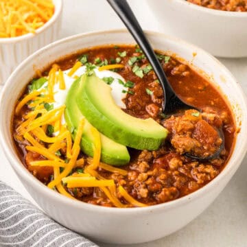 Square image of a white bowl filled with keto chili, with slices of avocado and shredded cheese on top.