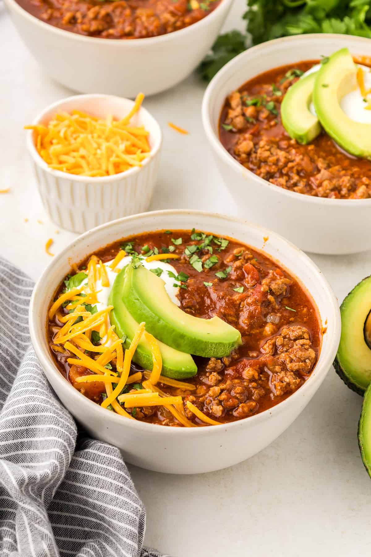 Three bowls of no bean chili on a light table top with a bowl of shredded cheese.