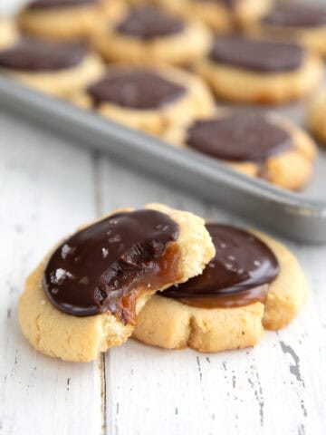 Two Keto Twix Cookies on a white wooden table with a tray of cookies in the background.