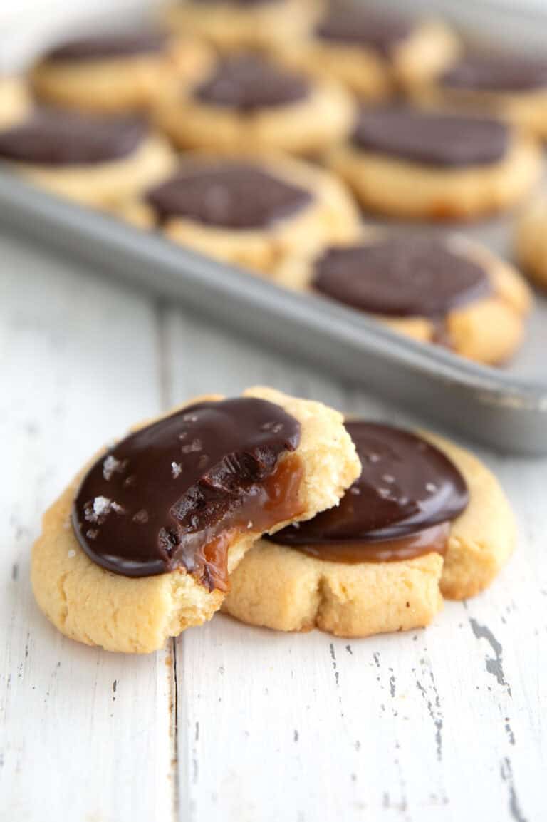 Two Keto Twix Cookies on a white wooden table with a tray of cookies in the background.