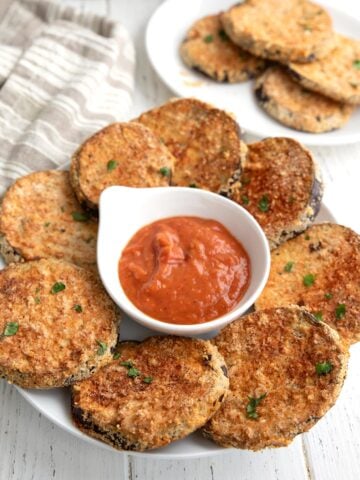 Pieces of Crispy Air Fryer Eggplant on a white plate with a bowl of marinara in the center.