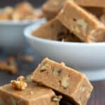 Titled image of two pieces of Keto Maple Walnut Fudge on a dark wooden table with a bowl of more fudge in the background.