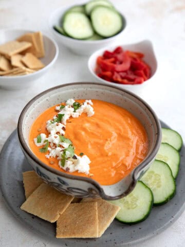 A bowl of Red Pepper Feta Dip on a gray plate with keto crackers and cucumber slices.