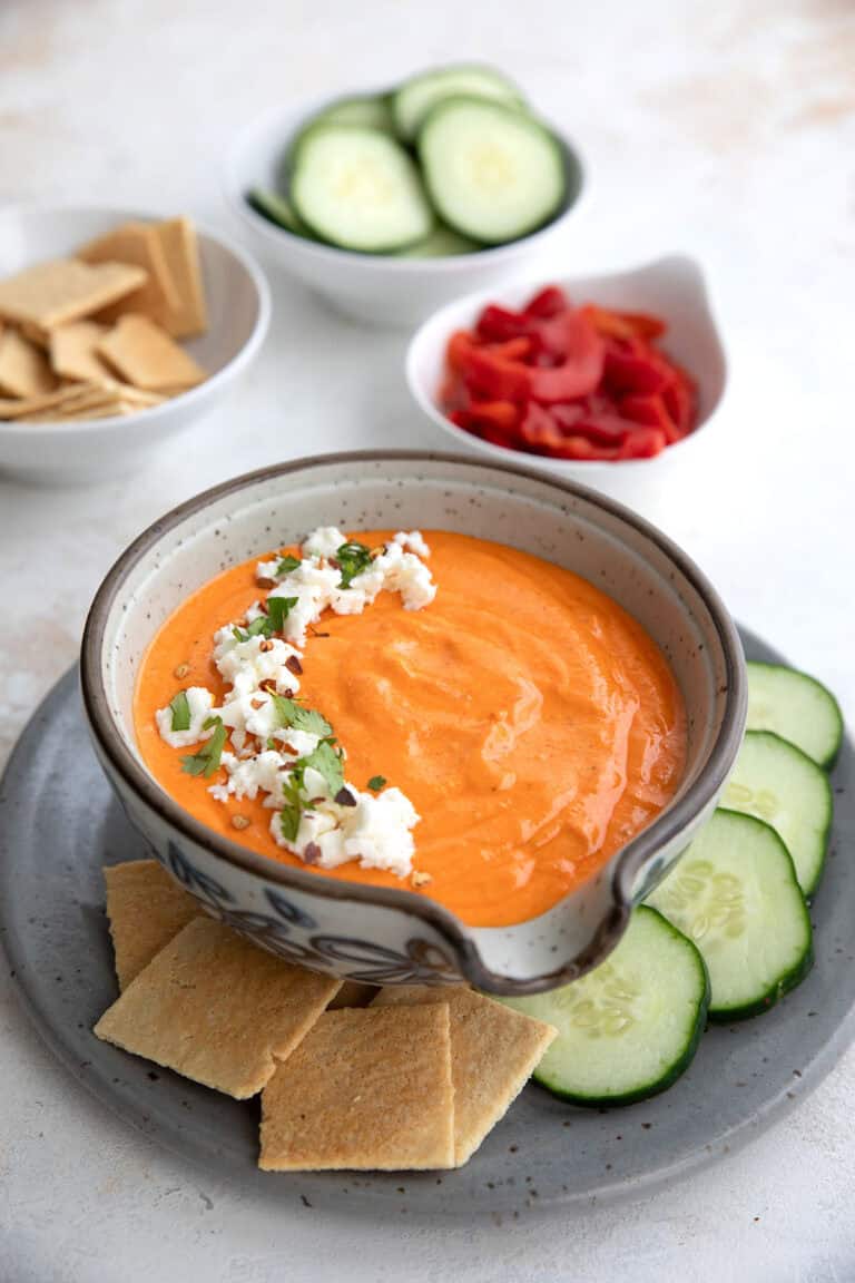 A bowl of Red Pepper Feta Dip on a gray plate with keto crackers and cucumber slices.