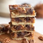 A stack of four Keto Pecan Pie Bars on a wooden cutting board with pecans strewn around.