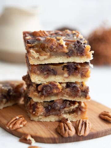 A stack of four Keto Pecan Pie Bars on a wooden cutting board with pecans strewn around.