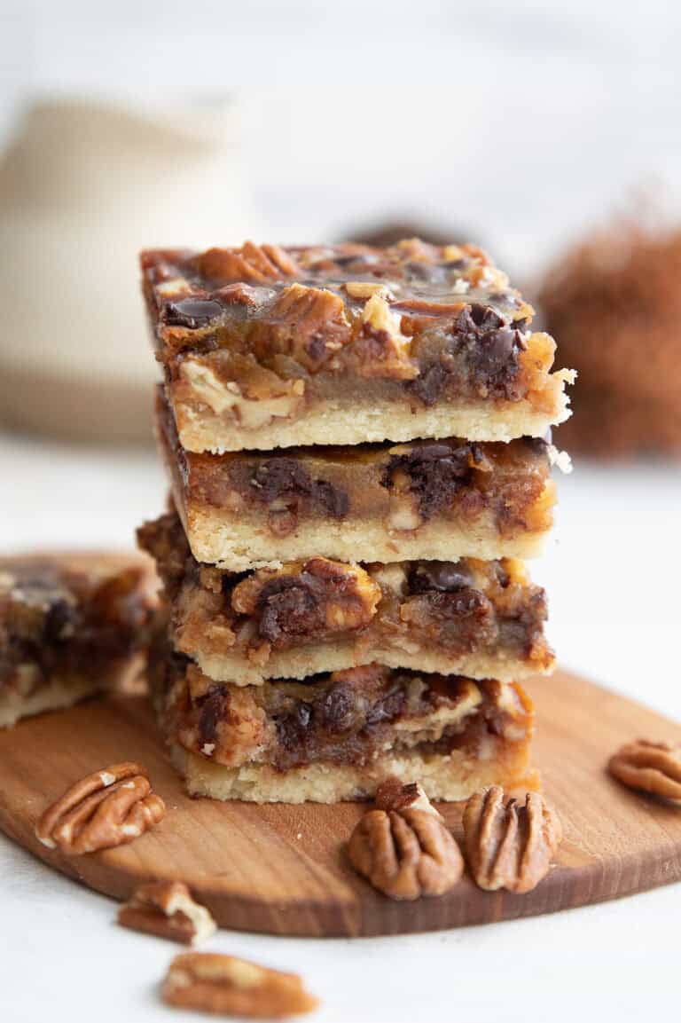 A stack of four Keto Pecan Pie Bars on a wooden cutting board with pecans strewn around.