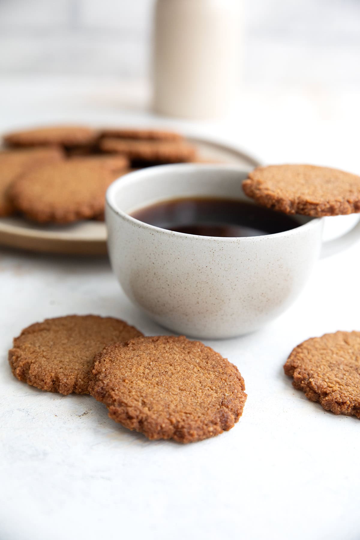 Keto Gingersnap Cookies on a white table with a cup of coffee. One cookie resting on the rim of the coffee.