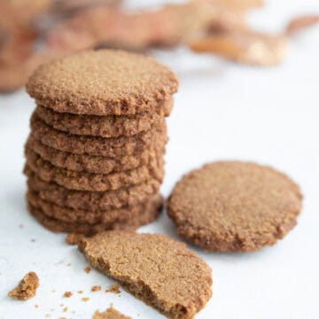 A stack of Keto Gingersnap Cookies on a white table with one broken in front, and a branch of golden brown leaves in the background.