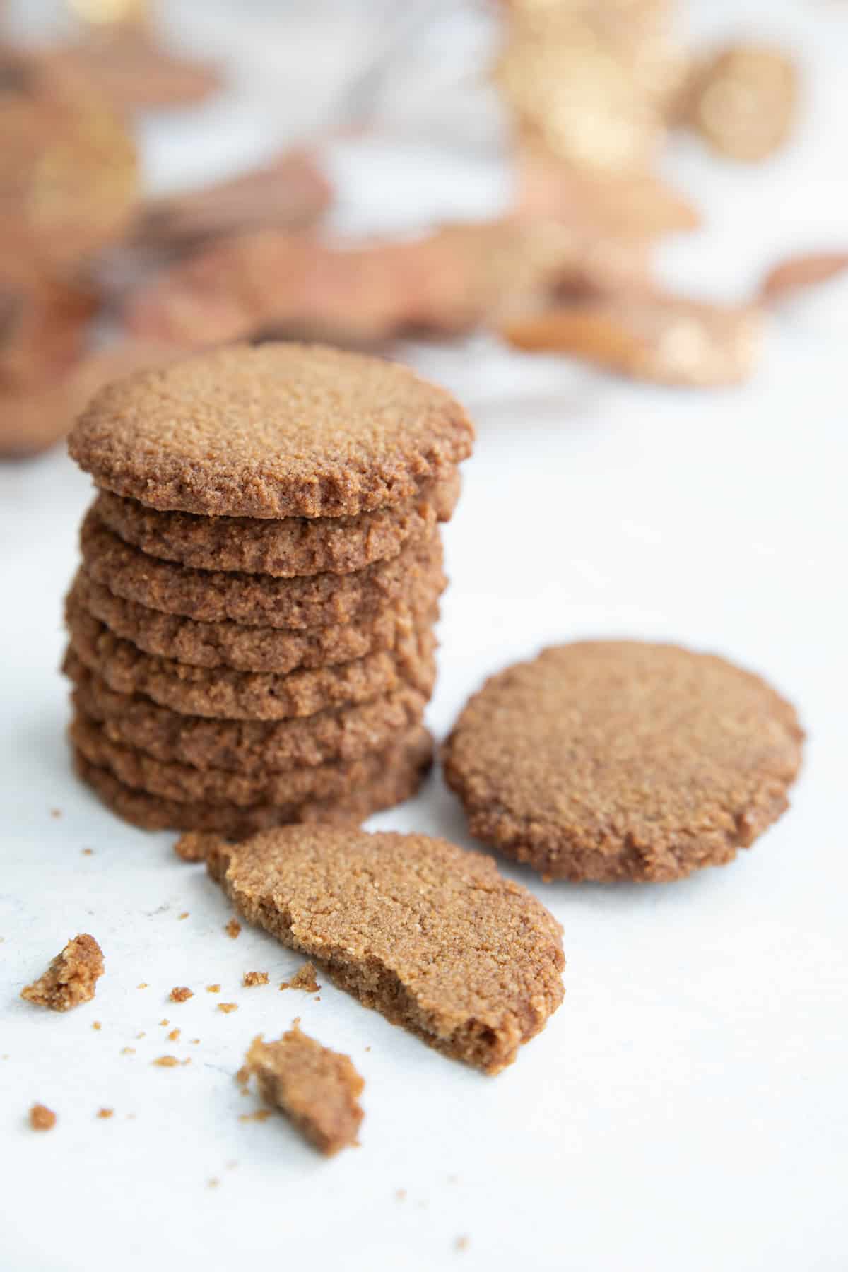 A stack of Keto Gingersnap Cookies on a white table with one broken in front, and a branch of golden brown leaves in the background.