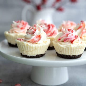 Keto White Chocolate Peppermint Cheesecakes on a white Cake stand with a vase of red winter berries in the background.