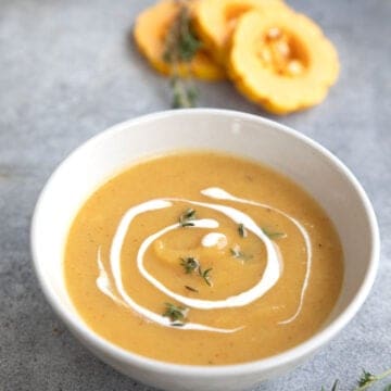 A white bowl filled with Roasted Delicata Squash Soup on a gray table, with rings of delicata squash in the background.